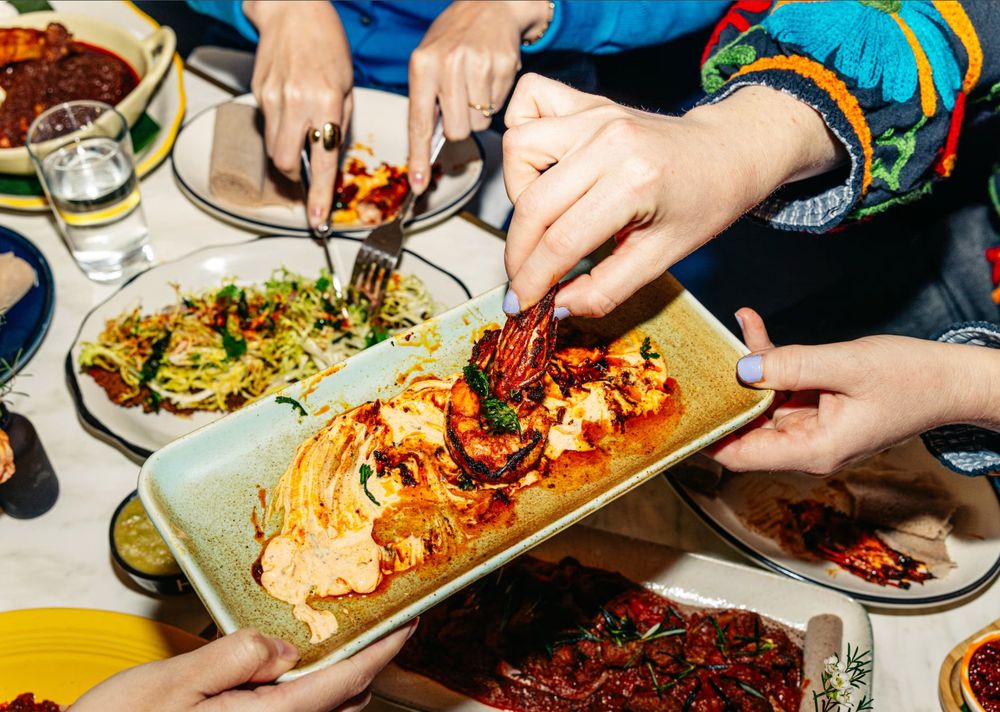 Hands sharing a grilled prawn dish over a spiced cream sauce