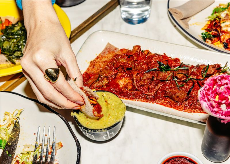 Hand dipping injera into avocado sauce beside a platter of saucy lamb